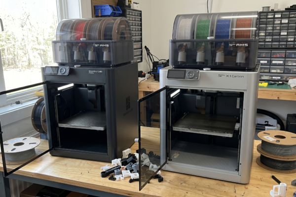 Bambu Lab P2S and X1 Carbon 3D printers side by side on a maker's workshop desk
