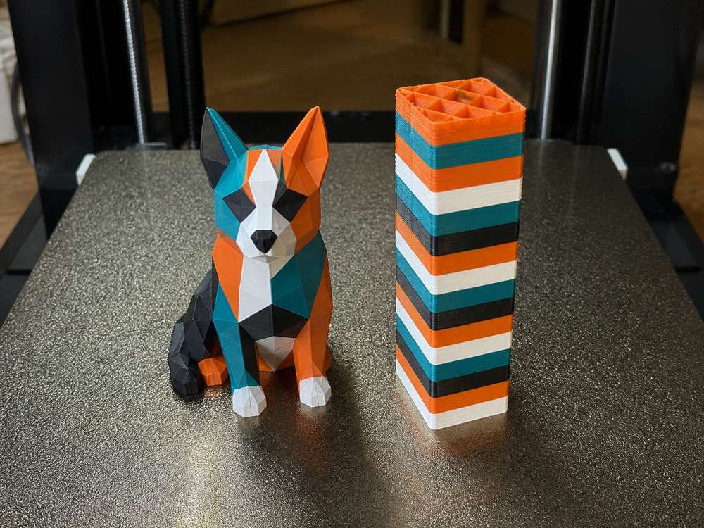 Multi-color 3D printed figurine next to a tall striped purge tower on a PEI build plate, showing AMS filament waste